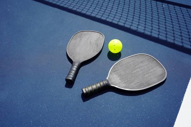 Two black textured pickleball paddles lying on a blue court with net shadows and a yellow ball