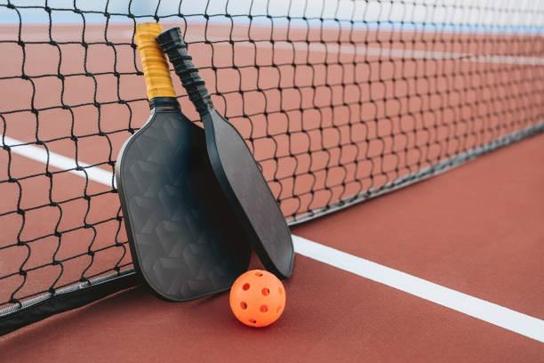 Two black pickleball paddles leaning against the net on a red court with an orange pickleball