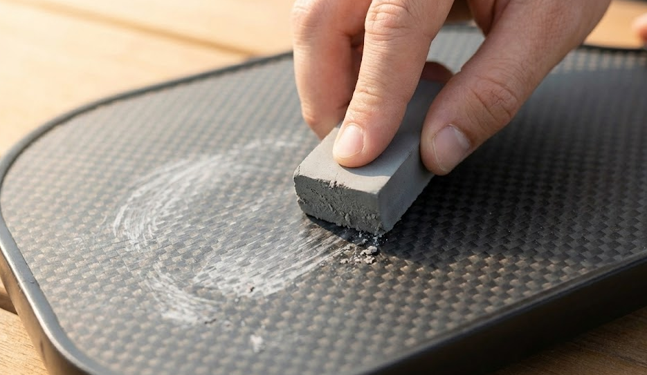 Removing plastic ball residue from the raw carbon fiber surface of pickleball paddles using a rubber eraser bar to restore spin potential.