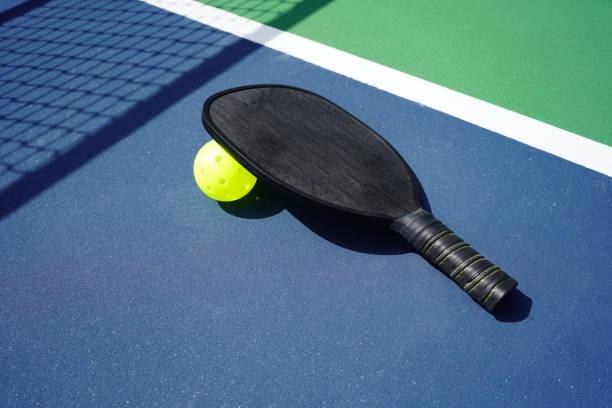 Sleek black pickleball paddle lying on a blue and green hard court surface with a yellow ball