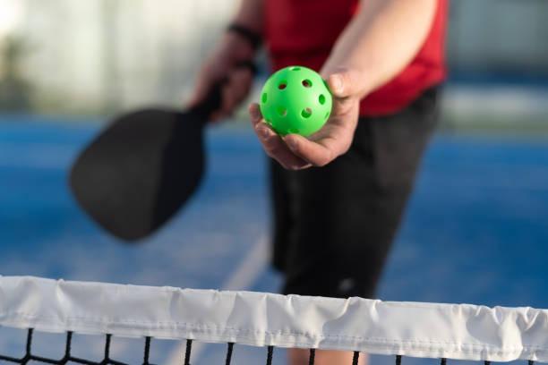 Close-up of player holding a green ball preparing to serve with a black pickleball paddle in the background
