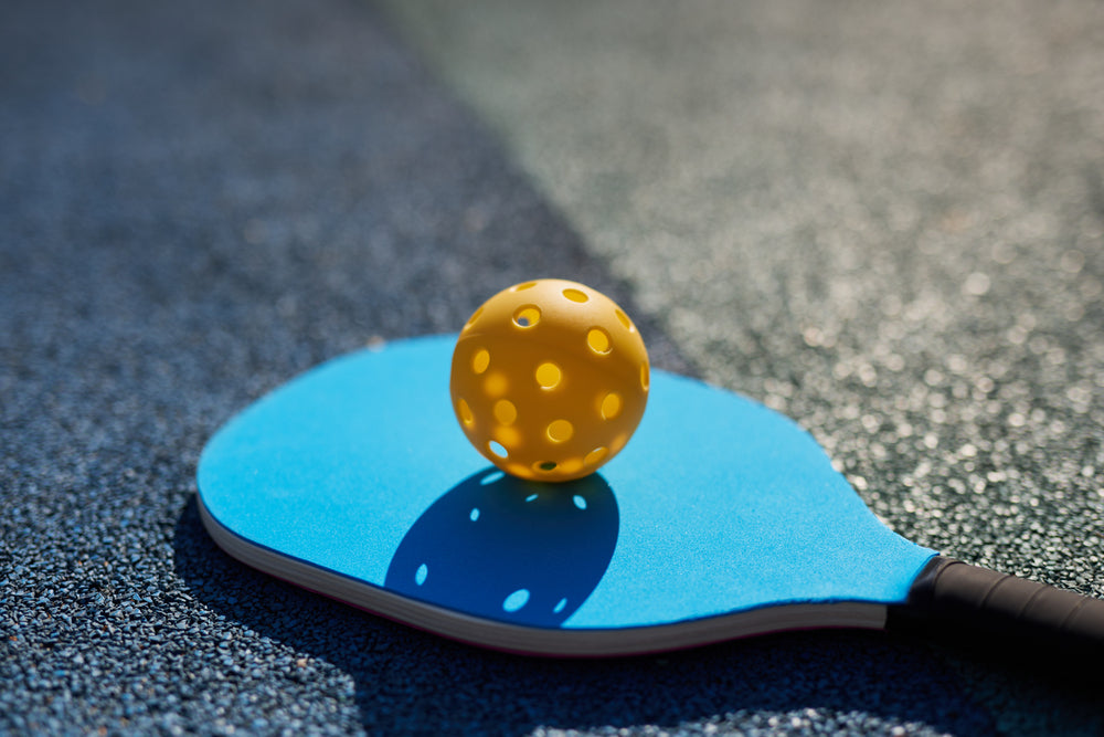 Yellow pickleball resting on a blue pickleball paddle on a court. Find great pickleball paddles.