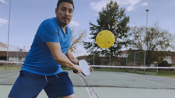 Man in blue preparing to hit a yellow pickleball with pickleball paddles on court.
