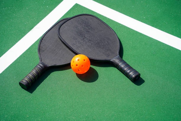 Two black pickleball paddles with orange ball on green court.