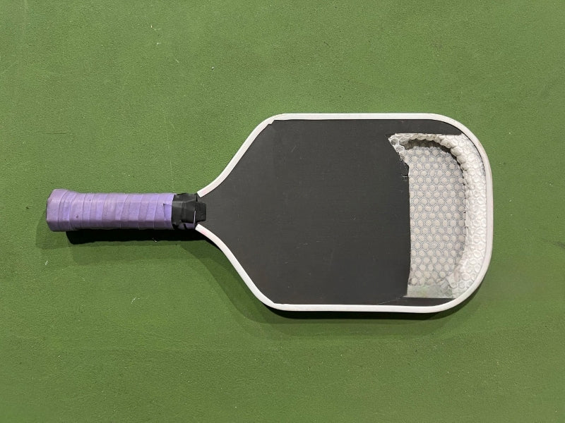 Black pickleball paddle with purple grip, revealing honeycomb core on a green court.