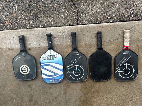 Collection of various used pickleball paddles resting on concrete, showcasing different brands.