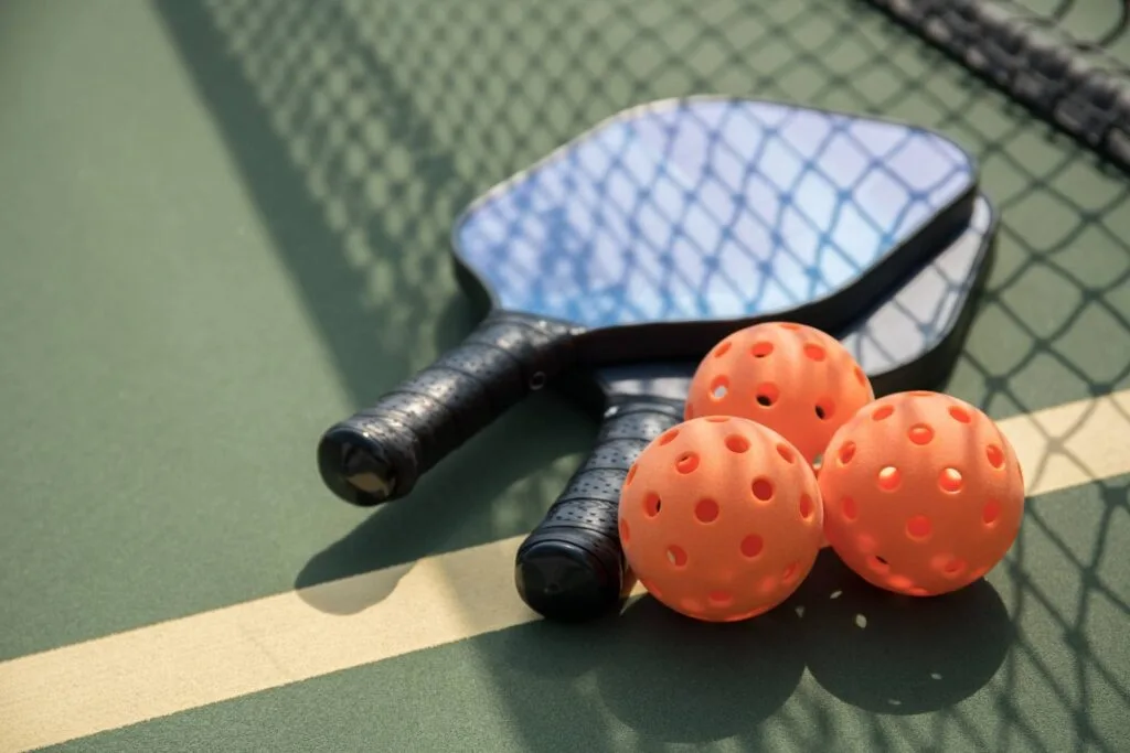 Two black pickleball paddles with three orange balls near the net on a court.