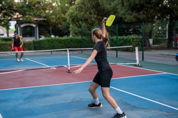 Woman swinging a yellow pickleball paddles on a court, playing with another person.