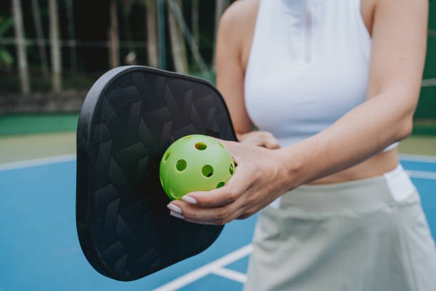 Close-up of hands holding a green pickleball and a black pickleball paddles on court.